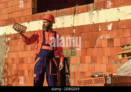 Backstein-Baumaterial für die Herstellung von Wänden. Leichter Backstein aus geschäumtem Tonaggregat. Element in der Maurerkonstruktion. Bauherr hat Backstein. Bärtiger Mann trägt auf der Baustelle eine harte Mütze. Stockfoto