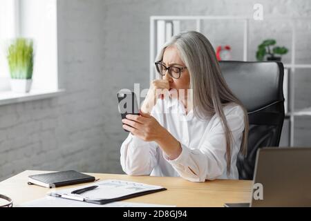 Verwirrt Senior grauhaarige schöne Frau hält Smartphone. Peinliche Geschäftsfrau hat Problem mit gebrochenen nicht funktioniert Handy. Empfangen Stockfoto