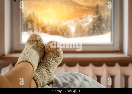 Frauenbeine in warmen Socken liegen. Fenster mit verschwommenem Blick auf den verschneiten Winter. Gemütliches Zuhause. Stockfoto