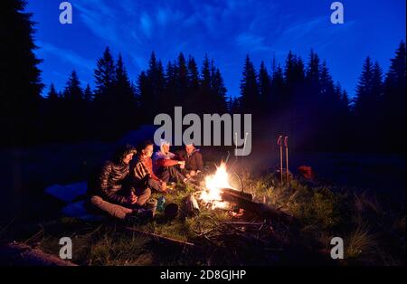 Abend Camping in der Nähe von Feuer, Fichtenwald im Hintergrund. Gruppe von vier Freunden sitzen zusammen am Lagerfeuer, genießen frische Luft in der Nähe Zelt mit Touristen Ausrüstung in der Nähe, dunkel entspannende Atmosphäre. Stockfoto