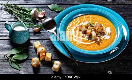 Cremig beruhigende Herbstgericht Kürbissuppe in einem blauen Teller mit Croutons und Kürbiskernen auf einem Deckel auf einem dunklen Holzhintergrund mit Sahne, Knoblauch, Ba Stockfoto