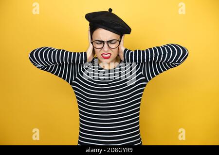 Junge schöne brünette Frau mit französischem Beret und Brille über gelbem Hintergrund, die an Kopfschmerzen verzweifelt und gestresst ist, weil Schmerzen und Stockfoto