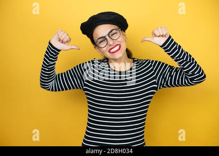 Junge, schöne Brunette-Frau mit französischem Beret und Brille über gelbem Hintergrund, die selbstsicher mit einem Lächeln im Gesicht aussieht und sich mit Finne zeigt Stockfoto