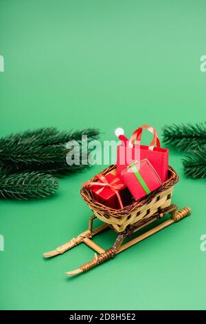 Geschenkboxen und Einkaufstasche mit weihnachtsmütze in Weide Korb auf dekorativem Schlitten bei Tannenzweigen auf Grün Stockfoto