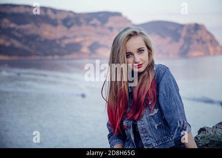 Junge blonde Mädchen mit hellen Funktionen lächeln auf dem Hintergrund des Meeres und der Berge in einem Jeansrock und Jacke. Hintergrund verschwommen für künstlerische Stockfoto