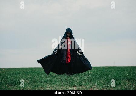 Junge Frau geht über Feld in Halloween Kostüm des Todes mit bemalten Skelett auf ihrem Körper vor Hintergrund Himmel. Halloween-Konzept. Zuckerschädel Stockfoto