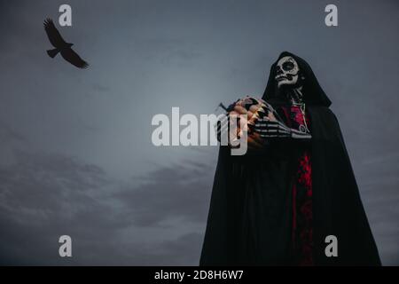 Junge Frau steht im Halloween Kostüm des Todes mit bemaltem Skelett auf ihrem Körper und Jack-o-Laterne vor dem Hintergrund von dunklem Himmel und fliegender Rabe. Stockfoto