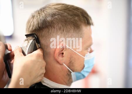 Junger Mann trägt schützende Gesichtsmaske bekommt Haarschnitt mit Haarschneider Im Friseur Stockfoto