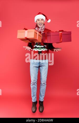 Positive Mann in santa hat springen in der Nähe Geschenke auf rot Hintergrund Stockfoto