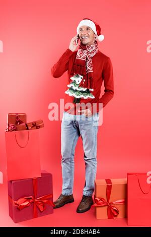 Lächelnder Mann in santa hat auf Smartphone in der Nähe Geschenk sprechen Boxen und Geschenke auf rotem Hintergrund Stockfoto