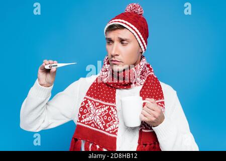 Kranker und verärgerter Mann in Strickmode, der Mütze hält und zeigt Thermometer auf blau isoliert Stockfoto