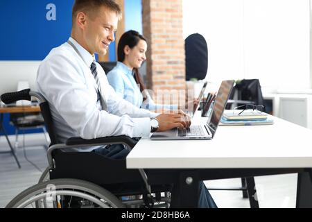 Ein junger lächelnder Mann im Rollstuhl arbeitet im Büro am Laptop Mit einer Kollegin Stockfoto