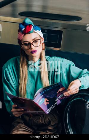 Stilvolle junge Frau in Turban und Brille Lesemagazin in Waschsalon Stockfoto