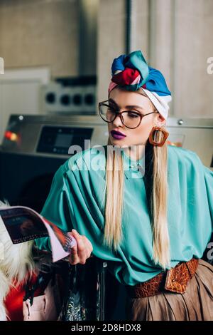 Junge und stilvolle Frau in Brille und Turban Lesemagazin Im Waschsalon Stockfoto