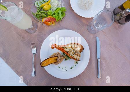 Gegrillter Hummer ist wunderschön auf einem Teller zusammen mit Beilage von Kartoffelpüree, gebratene Banane, Gemüse und Reis und ein Glas Wasser platziert Stockfoto
