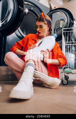 Junge Frau in Sonnenbrille hält Flasche mit Waschmittel und sitzen Auf dem Boden im Waschsalon Stockfoto