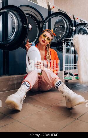 Junge Frau in Sonnenbrille hält Flasche mit Waschmittel und sitzen Auf dem Boden in der Nähe von Waschmaschinen und Wagen im Waschsalon Stockfoto