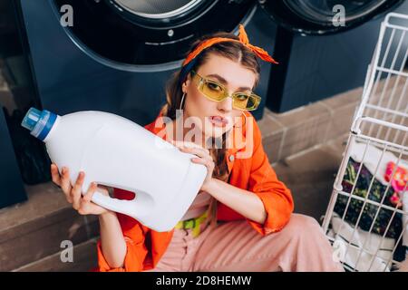 High-Winkel-Ansicht der jungen Frau auf dem Boden sitzen und Flasche mit Waschmittel im Waschsalon halten Stockfoto