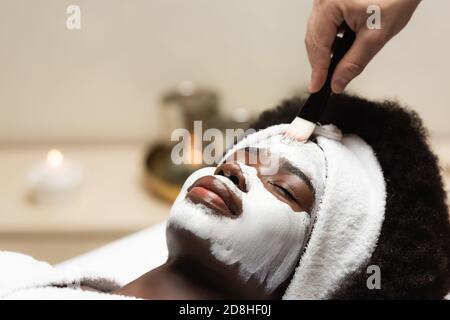 afroamerikanische Frau im Stirnband liegen in der Nähe Spa Therapeut Anwendung Gesichtsmaske auf Stirn auf verschwommenem Hintergrund Stockfoto