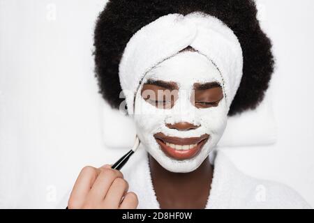 Draufsicht auf lächelnde afroamerikanische Frau mit Gesichtsmaske In der Nähe von Spa-Therapeuten mit Kosmetikbürste im Spa-Salon Stockfoto