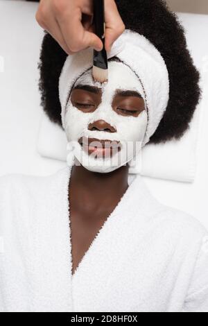 Draufsicht auf eine positive afroamerikanische Frau, die einen Bademantel trägt und in der Nähe des Spa-Therapeuten liegt, der im Spa-Salon Gesichtsmaske auf die Stirn trägt Stockfoto