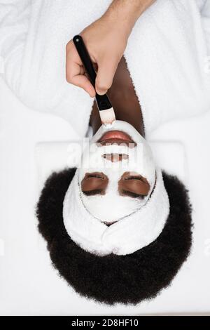 Draufsicht auf eine positive afroamerikanische Frau, die einen Bademantel trägt und in der Nähe des Spa-Therapeuten liegt, der im Spa-Salon Gesichtsmaske auf das Kinn aufträgt Stockfoto