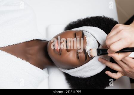 Spa Therapeut Anwendung Gesichtsmaske mit kosmetischen Pinsel auf der Stirn Der afroamerikanischen Frau trägt Frottee Mantel im Spa Salon Stockfoto