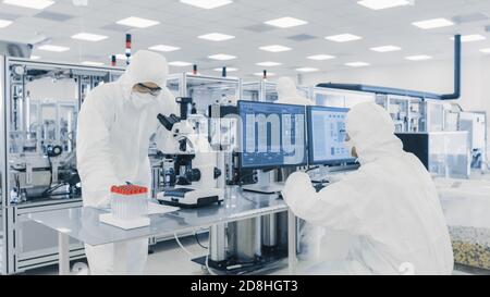 Team von Wissenschaftlern in sterilen Anzügen arbeiten mit Computern, Blick unter Mikroskop und moderne industrielle Maschinen im Labor Stockfoto