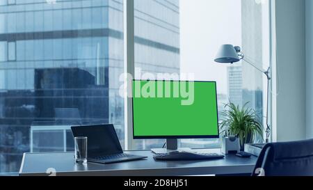 Aufnahme des Büroschreibtischs mit grünem Mock-up Bildschirm Personal Computer stehend darauf. Modernes, stilvolles Zimmer mit Blick auf das Geschäftsviertel der Großstadt. Stockfoto