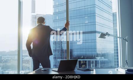 Aufnahme des selbstbewussten Geschäftsmannes in einem Anzug in seinem Büro, der gegen den Fensterrahmen schief und nachdenklich aus dem Fenster schaute. Großstadtgeschäft Stockfoto