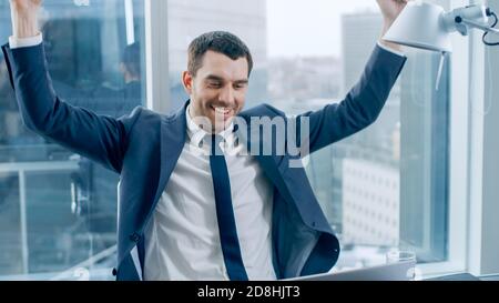 Erfolgreicher Geschäftsmann, der an seinem Schreibtisch sitzt, an einem Laptop arbeitet, gewinnt Big Deal und feiert das Erreichen des Ziels, die gewinnende Geste. Im großen Fenster Stockfoto