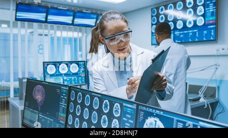 Senior Medical Scientist berät weibliche Lehrling mit Clipboard, Er arbeitet mit CT Brain Scan Bilder auf einem Personal Computer im Labor Stockfoto