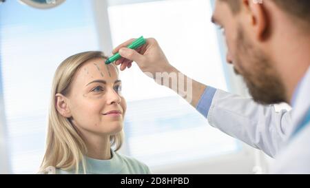 Over the Shoulder Shot eines plastischen Kosmetikchirurgen zeichnet Linien auf ein Gesicht eines schönen weiblichen Modells für die Facelift-Operation. Stockfoto