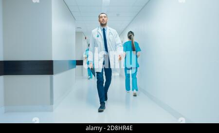 Determined Handsome Doctor trägt weißen Mantel mit Stethoskop geht durch Krankenhaus Flur. Moderne helle Klinik mit professionellem Personal. Stockfoto