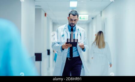 Determined Handsome Doctor nutzt Digital Tablet Computer beim Gehen durch Krankenhaus Flur. Moderne helle Klinik mit professionellem Personal. Stockfoto