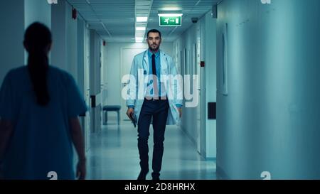 Determined Handsome Doctor trägt weißen Mantel mit Stethoskop geht durch Krankenhaus Flur. Moderne helle Klinik mit professionellem Personal. Stockfoto