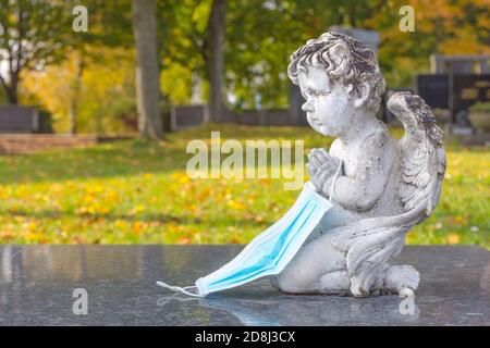 Opfer von Coronavirus Covid 19 Konzeptfoto mit Skulptur eines kleinen Engels, der eine schützende medizinische Maske auf einem Grab auf dem Friedhof hält. Selektive Stockfoto