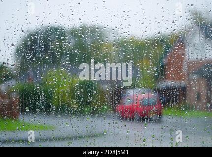 Verschwommener Blick durch nasses Fenster mit Regentropfen zu rotem Auto Und rote Backsteinhäuser mit Bäumen im Hintergrund Stockfoto