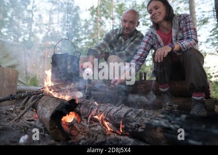 Zufriedene, liebliche Freunde braten eine köstliche Marshmallow über einem Feuer, über dessen Flamme ein Wasserkocher mit einem heißen Getränk hängt. Stockfoto