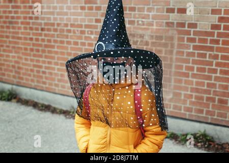 Frohe Halloween. Kind Mädchen in Hexenhut und Gesicht Schutzmaske gehen zur Schule. Schulschüler feiern Halloween-Urlaub. Eine neue Normalität während c Stockfoto