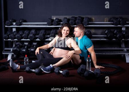 Paar kaukasischen starken und muskulösen Mann und sportliche schwangere Frau Entspannen Sie sich, während Sie auf dem Boden in der Nähe der Fitnessgeräte im Fitnessraum sitzen Für Cross-Training Stockfoto