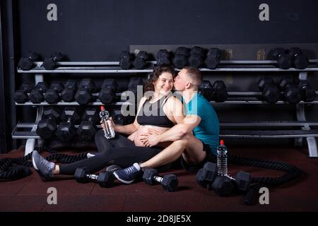 Paar kaukasischen starken und muskulösen Mann und sportliche schwangere Frau Entspannen Sie sich, während Sie auf dem Boden in der Nähe der Fitnessgeräte im Fitnessraum sitzen Für Cross-Training Stockfoto