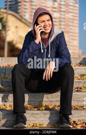 Lächelnder, fröhlicher Teenager-Junge, der auf dem Mobiltelefon spricht und auf dem er sitzt Treppen im Freien in der Stadt Stockfoto