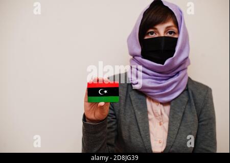 Portrait der jungen muslimischen Frau trägt formelle Kleidung, schützen Gesichtsmaske und Hijab Kopftuch, halten Libyen Flagge Karte vor isoliertem Hintergrund. Coronavi Stockfoto