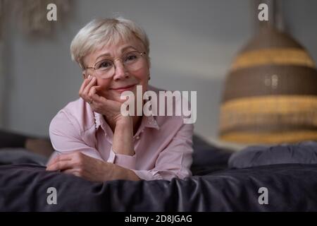 Ältere kaukasische Frau, die sich auf dem Bett entspannt und die Kamera anlächelt Stockfoto