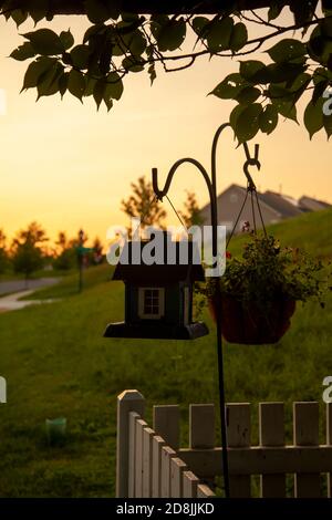 Ein abstraktes Konzeptbild, das bei Sonnenuntergang aufgenommen wurde und einen Stand-Alone-Metallaufhänger mit einem Vogelhaus und einem Blumentopf zeigt. Das Rack ist platziert Stockfoto