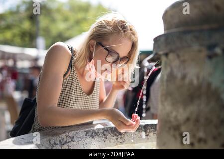 Durstige junge Casual cucasian Frau trägt medizinische Gesichtsmaske Trinkwasser aus öffentlichen Stadtbrunnen an einem heißen Sommertag. Neue soziale Normen während Stockfoto