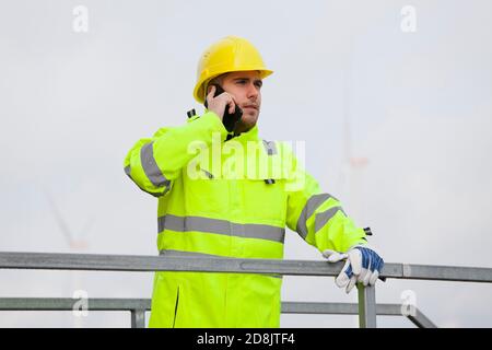 Junger Ingenieur oder Arbeiter, der vor dem Telefon spricht Von Windenergieanlagen im unscharfen Hintergrund Stockfoto