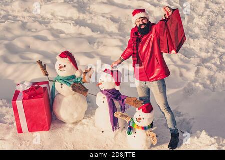 Aufgeregt Mann mit lustigen Schneemann in stilvollen Hut und Schal auf schneebedeckten Feld. Happy Winter Schneemann Familie mit Geschenk. Mutter Schnee-Frau, Vater Schnee-Mann und Stockfoto