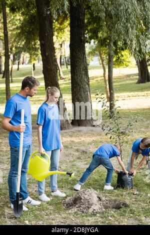 Kinder tragen jungen Baum, während die Eltern mit Schaufel und Gießkanne stehen, Ökologie Konzept Stockfoto